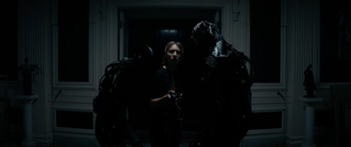A woman stands between two shadowy, armored figures in a dark room in a still from the gothic horror thriller Recluse.